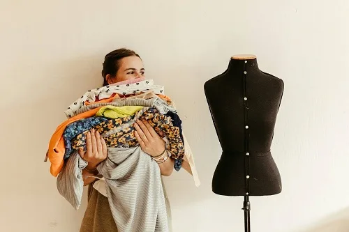 A fashion designer holding a pile of clothes while standing next to and looking at a black mannequin