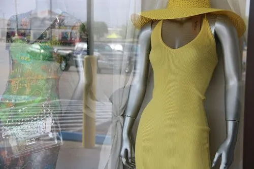A mannequin behind glass in a shop window wearing a yellow dress